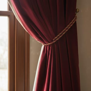 May include: Burgundy-colored curtains with a gold and red rope tieback. The curtains are draped and hang in front of a window with a wooden frame. The tieback is wrapped around the curtains to hold them open.