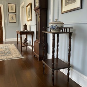 May include: A dark brown, antique plant stand with a two-tiered design. The stand features turned legs and a small top shelf. A blue and white floral ceramic bowl with a lid sits on the top shelf. The stand is in a hallway with a grandfather clock.