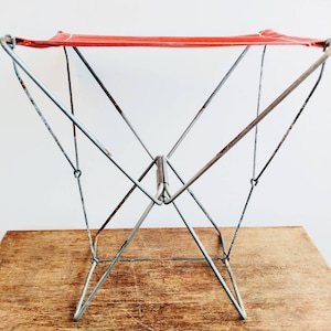 May include: A red canvas folding stool with a metal frame. The stool is made of thin metal rods that are connected together to form a triangular shape. The stool is folded up and sitting on a wooden surface.