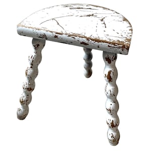 May include: A small, white, distressed wooden stool with a half-moon shaped seat. The seat and legs show signs of wear, with exposed wood peeking through the white paint. The legs are turned with a bobbin design.