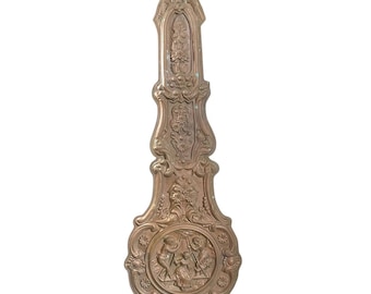 May include: A gold-toned metal pendulum with intricate floral and scrollwork designs. The pendulum features a central circular panel with a scene of three figures.