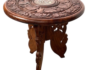 May include: A round, intricately carved wooden table with a floral design. The table has a small, inlaid circular top and three legs that fold for easy storage.