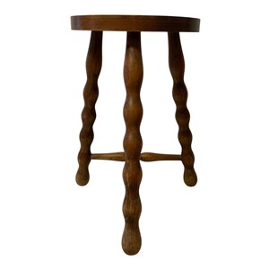 K&ouml;nnte beinhalten: Ein kleiner, dreibeiniger Holzschemel mit rundem Sitz. Die Beine sind aufwendig mit einem gedrehten, seilartigen Design geschnitzt. Der Hocker ist aus warmem, mittelget&ouml;ntem Holz gefertigt und zeigt die nat&uuml;rliche Maserung.