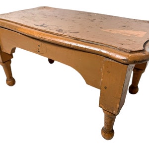 Puede incluir: Un pequeño taburete de madera con un acabado marrón claro. El taburete tiene una parte superior curva y patas torneadas.