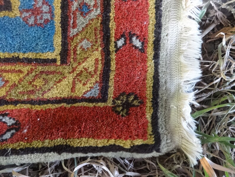 May include: Close-up of the edge of a rug with a red, gold, and blue pattern. The rug has a fringe border and is made of a soft, woven material.