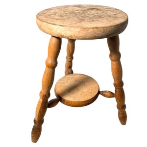 May include: A wooden stool with a round seat and three turned legs. The stool is made of light brown wood and has a natural finish.