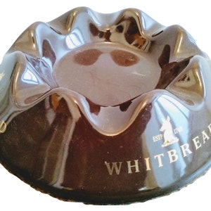 May include: A brown ceramic ashtray with a gold inscription that reads "WHITBREAD ESTP. 1746" and a stylized rabbit logo.