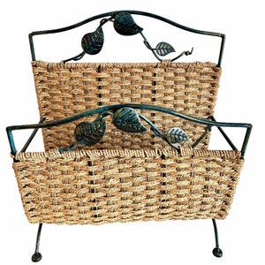 May include: A two-tiered magazine rack with a woven wicker design and metal frame. The frame is decorated with leafy vines and has a dark green finish.