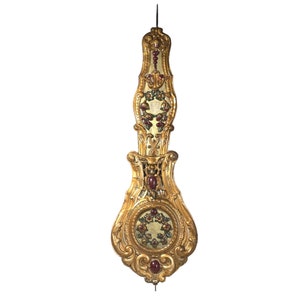 May include: A gold-coloured pendulum clock with intricate carvings and red accents. The clock face is surrounded by a wreath of green leaves and red berries.
