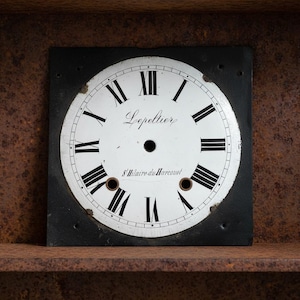 May include: A square clock face with a white dial and black Roman numerals. The clock face has the name "Lepeltier" and "S'Hilaire du Harcouet" printed on it. The clock face is set in a black square frame.