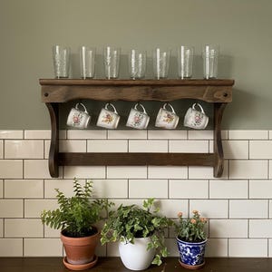 May include: A dark wooden shelf holds six etched drinking glasses and six floral teacups. The shelf is mounted above white tiles and potted plants, creating a vintage kitchen aesthetic. The shelf is approximately 60 cm wide.