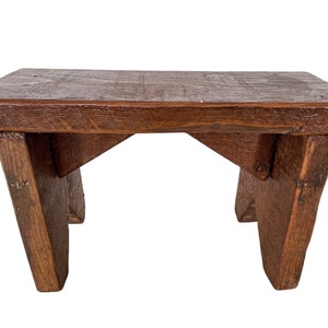 Puede incluir: Un taburete de madera rústico con un tinte marrón oscuro. El taburete tiene un diseño sencillo con una parte superior rectangular y cuatro patas.