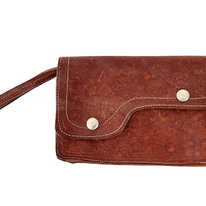 May include: A brown leather clutch with a flap closure and a short handle. The clutch has two silver metal snaps.