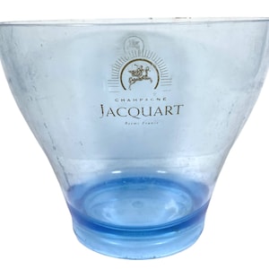 May include: A clear blue plastic champagne ice bucket with the Jacquart Champagne logo printed on the side. The logo features a golden sunburst with a horse and rider in the centre. The text "Champagne Jacquart Reims France" is printed below the logo.