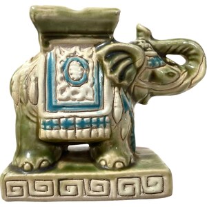May include: A green ceramic elephant figurine with blue and white details. The elephant is standing on a green base with a decorative border. The elephant has a saddle and is holding its trunk up.