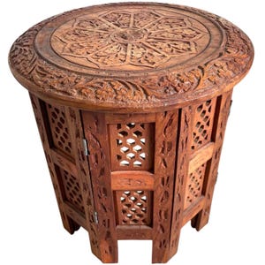 Może przedstawiać: Okrągły, bogato rzeźbiony drewniany stolik z ozdobnym blatem i ażurowymi bokami. Stolik wykonany jest z ciemnobrązowego drewna i ma tradycyjny wzór.