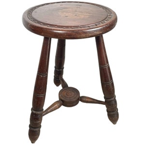 May include: A dark brown wooden stool with a round seat and three legs. The seat has a carved design around the edge.