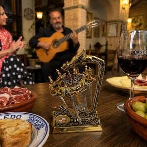 Peut inclure: Un porte-fourchettes à tapas décoratif en or et argent avec un motif de dragon. Le support est sur une table en bois avec un verre de vin rouge, des olives et de la charcuterie. Une personne joue de la guitare en arrière-plan.