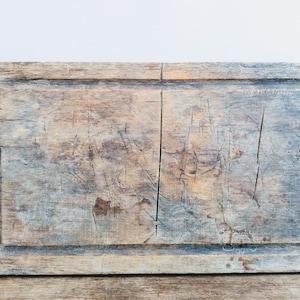 Op de afbeelding: Een verweerd houten snijplank met een rechthoekige vorm en een licht onregelmatige rand. De plank is lichtbruin van kleur en vertoont tekenen van gebruik met talrijke krassen en markeringen.