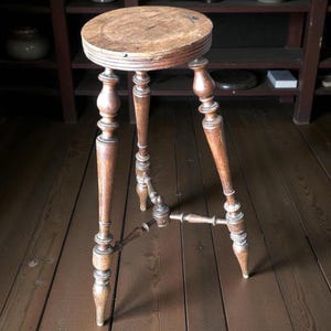 May include: A vintage wooden stool with a round seat and three ornate legs. The stool is made of light brown wood and has a distressed appearance, suggesting age and use. The legs are connected by a lower support.