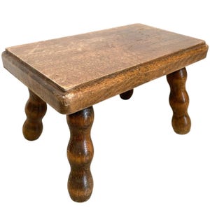 Könnte beinhalten: Ein Holzhocker mit einer rechteckigen Oberseite und vier gedrehten Beinen. Der Hocker ist aus dunkelbraunem Holz gefertigt und hat ein rustikales Aussehen.