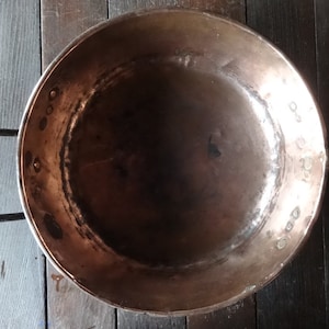 Könnte beinhalten: Ein großer Kupfertopf mit zwei schwarzen Metallgriffen. Der Topf ist leer und hat ein abgenutztes, altes Aussehen.