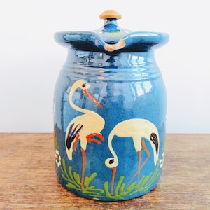 Peut inclure: Pichet en céramique bleu avec un couvercle en bois et des cigognes blanches peintes avec des becs et des pattes orange. Le pichet est recouvert d'un glaçage bleu et décoré de feuilles vertes et de fleurs blanches.