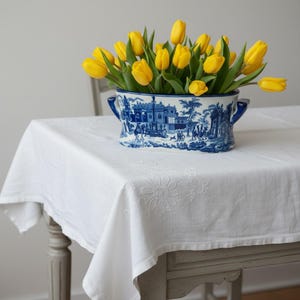 May include: A white tablecloth with embroidered floral details draped over a table, topped with an oval ceramic planter featuring a blue and white transferware pattern of architectural scenes and figures, filled with bright yellow tulips.