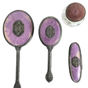 May include: A set of four vintage vanity items with silver-toned handles and purple fabric accents. The set includes two hand mirrors, a small compact, and a hairbrush.