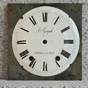 May include: A vintage clock face with a white background and black Roman numerals. The clock face has the text "A. Grayilu" and "à Villedieu-les-Poêles" in black script. The clock face is mounted on a square, gray metal plate.