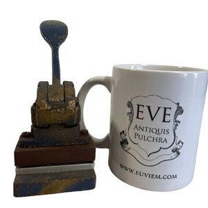 May include: A vintage blue and gold metal hand press with a wooden base and a white ceramic mug with the text "EVE ANTIQUIS PULCHRA www.euviem.com".