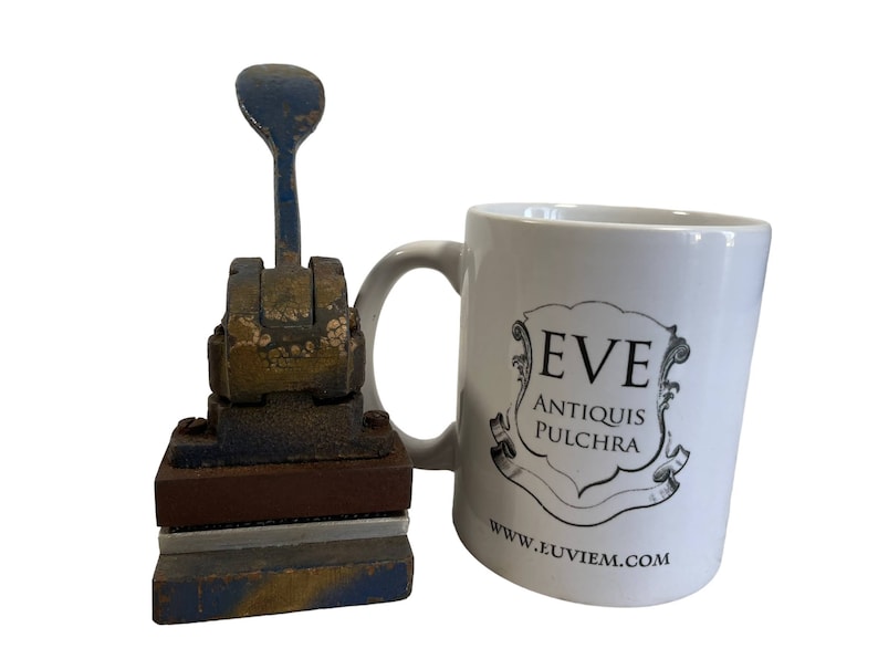 May include: A vintage blue and gold metal hand press with a wooden base and a white ceramic mug with the text "EVE ANTIQUIS PULCHRA www.euviem.com".