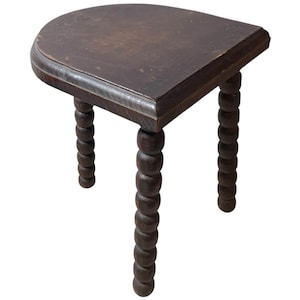 May include: A small, dark brown wooden stool with a semi-circular top and three turned legs. The legs are made of stacked, rounded wooden beads. The stool has a simple, rustic design and is likely vintage.