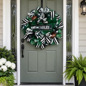 May include: A decorative wreath featuring green mesh, black and white striped ribbons, and football-themed accents. The center includes a rectangular sign with the text EAGLES and the team logo, surrounded by miniature brown footballs and green fabric.