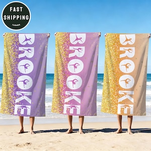 May include: Three personalized beach towels with the name "Brooke" in white, featuring a gymnast silhouette. The towels are in shades of purple, pink, and tan, with a gold glitter design on the left side. The towels are displayed on a beach.