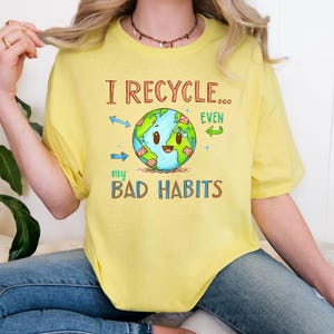 Può includere: T-shirt giallo chiaro con la scritta "I RECYCLE... EVEN my BAD HABITS". Il disegno presenta un pianeta sorridente circondato da simboli di riciclaggio. Il testo e la grafica sono in stile cartoon.