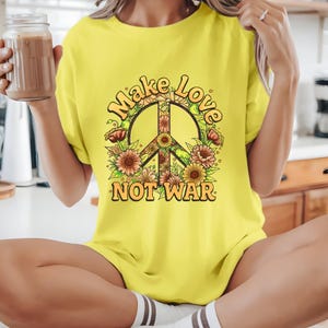 Puede incluir: Una camiseta amarilla con un diseño de símbolo de la paz y las palabras "Make Love Not War". El símbolo de la paz está lleno de patrones florales y rodeado de flores. El texto está en una fuente retro.