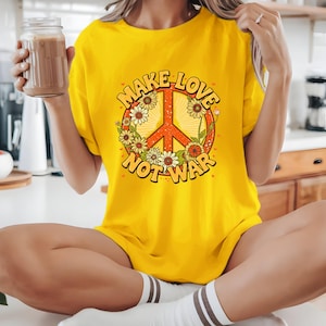 Puede incluir: Camiseta amarilla brillante con un diseño de signo de la paz. El gráfico incluye un símbolo de la paz naranja, las palabras "MAKE LOVE NOT WAR" y elementos florales. La camiseta es de color amarillo liso, perfecta para el uso diario.