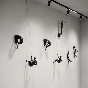 Könnte beinhalten: Schwarze Wandkunst mit Kletterfiguren an Seilen. Die Figuren befinden sich in verschiedenen Positionen, einige an der Wand befestigt, andere im Aufstieg. Die Kunst wird an einer weißen Wand ausgestellt.