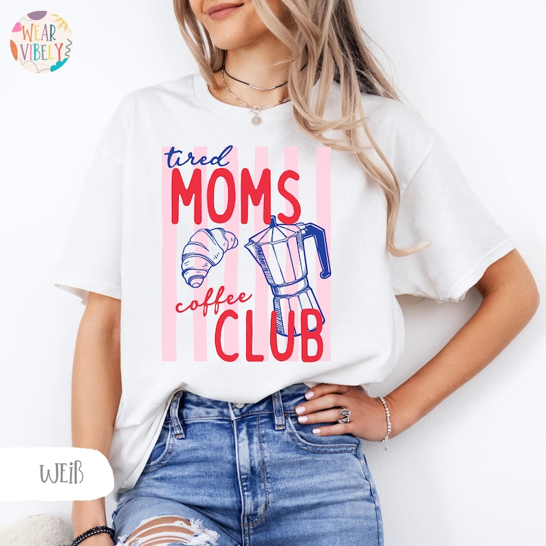 Pode incluir: T-shirt branco com a frase "tired MOMS coffee CLUB" em vermelho e azul, acompanhada por um croissant e um desenho de cafeteira. O fundo apresenta riscas verticais cor-de-rosa.