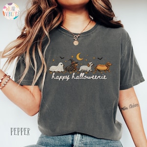 May include: A charcoal gray short-sleeve t-shirt featuring a graphic of four dachshunds in Halloween costumes, including a ghost, a witch, a mummy, and a hot dog. Above the dogs are a yellow crescent moon and bats. The text reads happy halloweenie.
