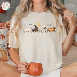May include: An ivory short-sleeve t-shirt featuring a graphic print of four dachshund dogs in Halloween costumes, including a ghost, a witch, a mummy, and a hot dog, set against a crescent moon, stars, and bats in a brown and orange color palette.