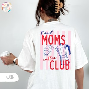 Pode incluir: Camiseta branca com um gr&aacute;fico nas costas que diz "tired MOMS coffee CLUB" em vermelho e azul. O gr&aacute;fico inclui um croissant e uma cafeteira. A camiseta tem um corte folgado.