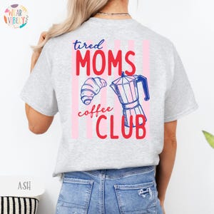 Pode incluir: T-shirt cinza claro com o texto "tired MOMS coffee CLUB" em vermelho e azul. O design inclui um croissant e uma cafeteira. Listras verticais rosa est&atilde;o atr&aacute;s do texto.