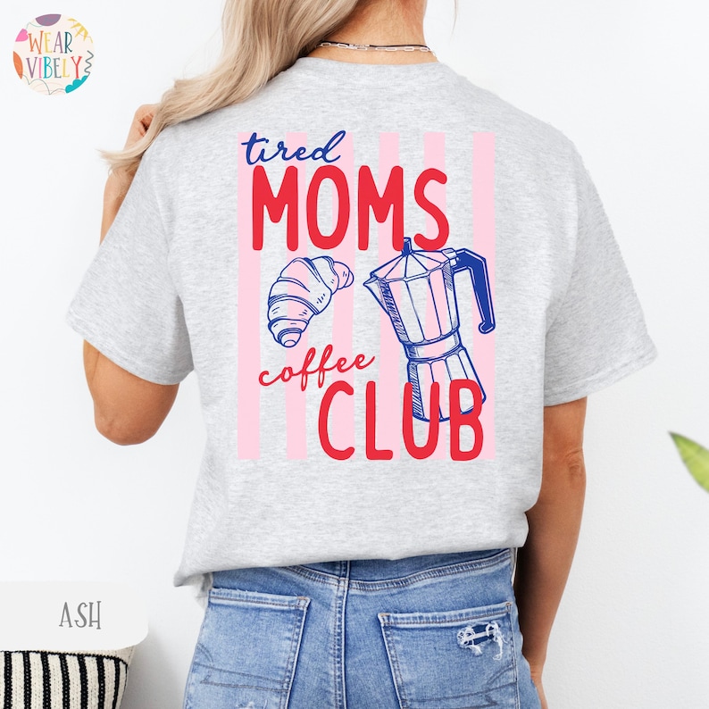 Pode incluir: T-shirt cinza claro com o texto "tired MOMS coffee CLUB" em vermelho e azul. O design inclui um croissant e uma cafeteira. Listras verticais rosa est&atilde;o atr&aacute;s do texto.