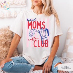 Pode incluir: Camiseta creme com a frase "tired MOMS coffee CLUB" em vermelho e rosa, juntamente com um croissant e um gr&aacute;fico de cafeteira. A camisa tem um corte folgado e mangas curtas.