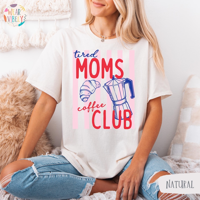 Pode incluir: Camiseta creme com a frase "tired MOMS coffee CLUB" em vermelho e rosa, juntamente com um croissant e um gr&aacute;fico de cafeteira. A camisa tem um corte folgado e mangas curtas.
