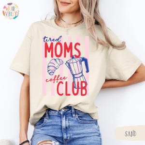 Pode incluir: T-shirt cor de areia com a frase "tired MOMS coffee CLUB" em vermelho e rosa. O design inclui um croissant e uma cafeteira. A camisa &eacute; uma pe&ccedil;a de roupa casual.