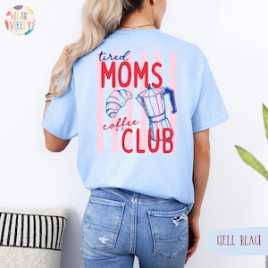 Pode incluir: Camiseta azul claro com o texto "tired MOMS coffee CLUB" em vermelho e azul. O design inclui um croissant e uma cafeteira. A parte de tr&aacute;s da camiseta tem listras verticais rosa e brancas.