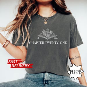May include: Dark gray t-shirt with white text and graphic. The graphic is an open book with flowers and the text "est. 2004 CHAPTER TWENTY-ONE". The shirt has a relaxed fit.